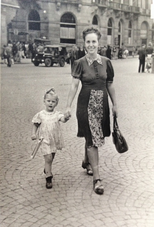 Juf Sissy loopt als 2-jarig meisje aan de hand van haar moeder op de Amsterdamse Dam op 7 mei 1945. Ze heeft een ratel in haar hand, speelgoed waar je herrie mee kan maken. 