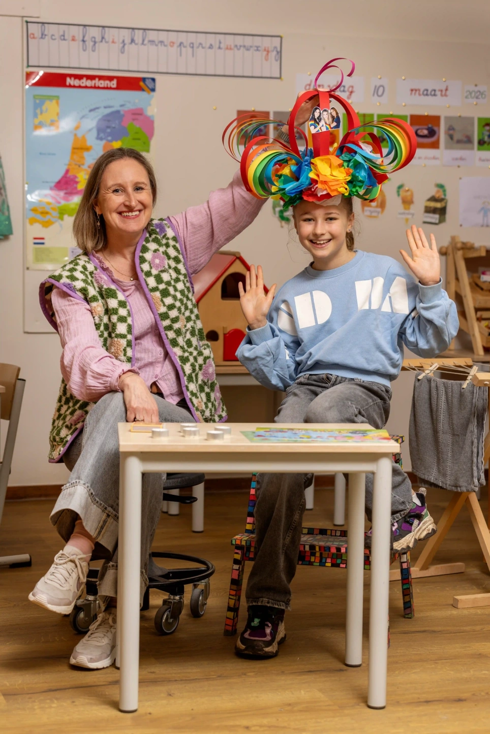 Portret van kleuterjuf Linda Rutjens samen met leerling Livia die een regenboog verjaardagsmuts op haar hoofd heeft - beeld bij aob.nl/onderwijsblad bij artikel over verjaardagsmutsen die lang bewaard worden