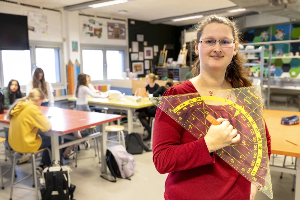 Portret van docent Maya Laan waarbij zij een grote geodriekhoek vasthoudt en in haar wiskundelokaal staat - beeld bij AOb.nl/onderwijsblad over docenten die twee vakken geven