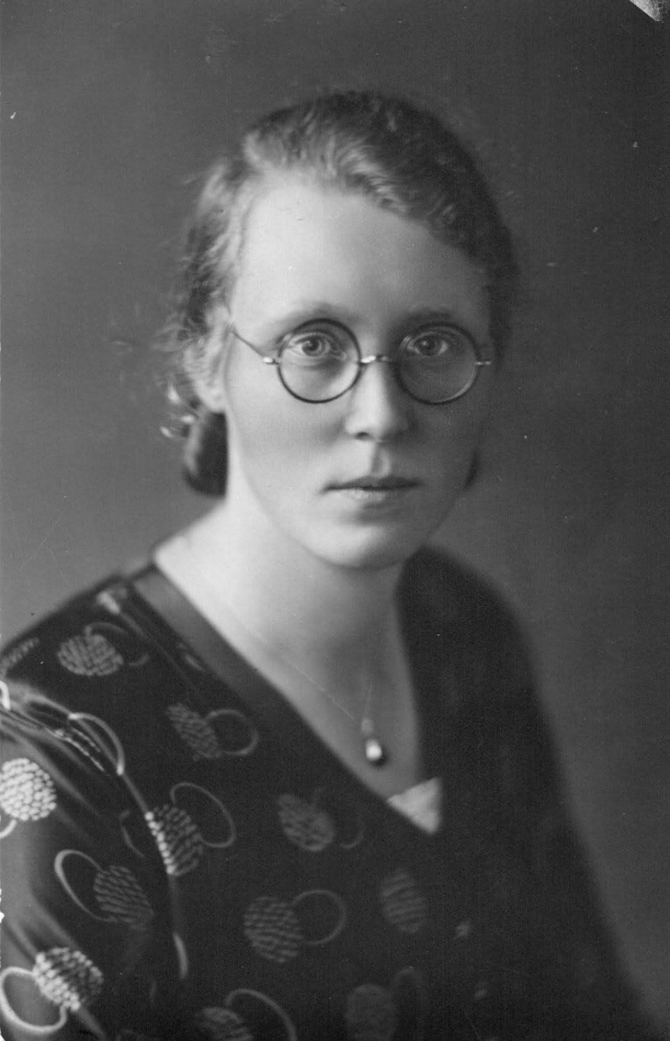 Zwart-witportret van een jonge vrouw met ronde bril, opgestoken haar en een serieuze blik. Ze draagt een donkere blouse met een patroon van cirkelvormige motieven en een eenvoudige ketting met een hangertje. De achtergrond is effen en vaag, wat de aandacht volledig op haar gezicht richt.