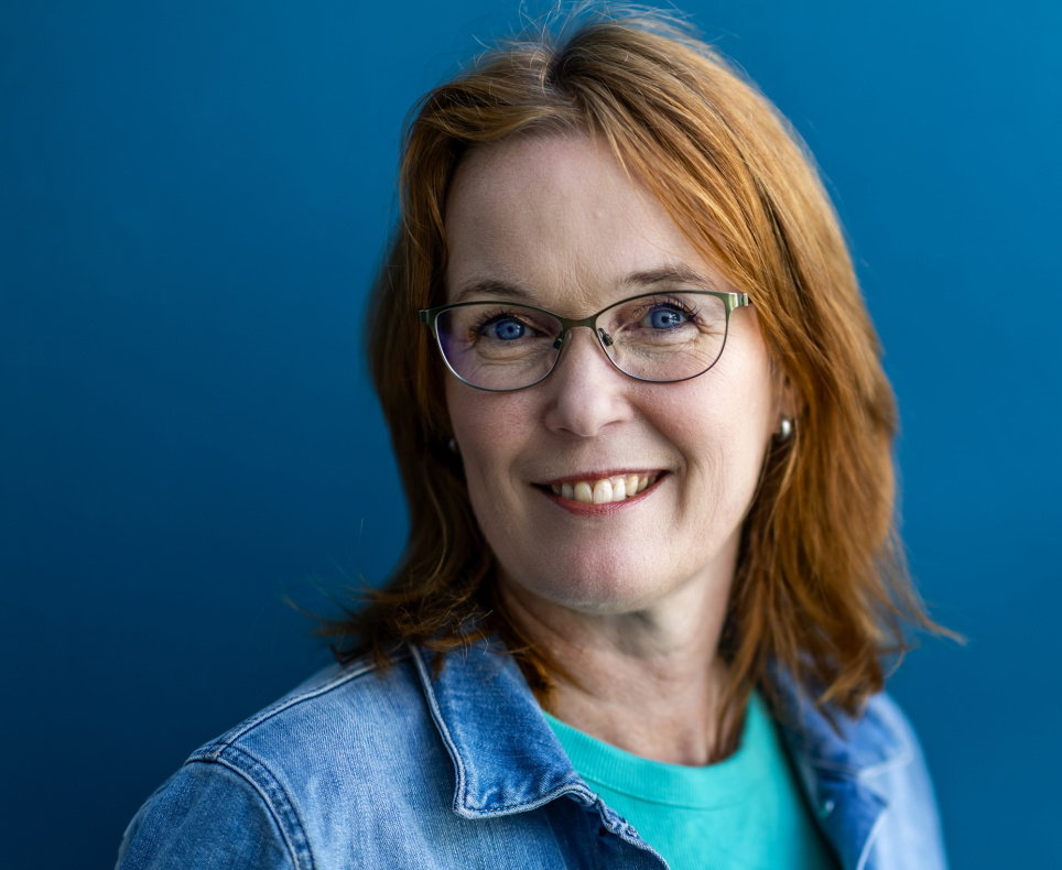 Portret van een lachende vrouw met halflang roodbruin haar en een bril. Ze draagt een turquoise shirt onder een licht spijkerjasje. De achtergrond is effen blauw, wat haar gezicht en kleding laat opvallen. De sfeer van de foto is vriendelijk en professioneel.