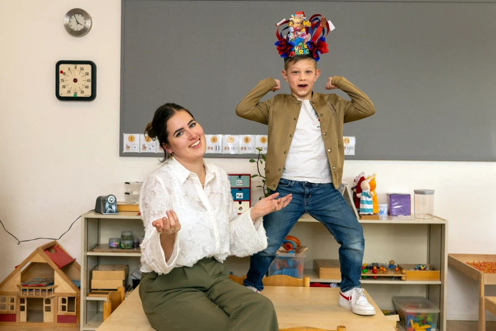 Portret van kleuterjuf Demi Kieft samen met Noud Stam uit groep 3 die op en tafel naast haar staat met zijn verjaardagsmuts van Super Mario op die Kieft voor hem maakte in de kleuterklas - beeld bij een artikel op aob.nl/onderwijsblad over verjaardagsmutsen