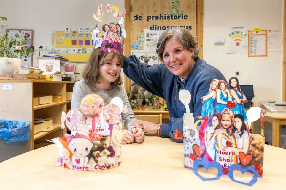 Portret van kleuterdocent Jacomine Wasser met Shae Pelgrom die nu in groep 8 zit maar al haar verjaardagsmutsen van Wasser heeft bewaard en ze toont op de tafel voor hen - beeld bij artikel op aob.nl/onderwijsblad over verjaardagsmutsen