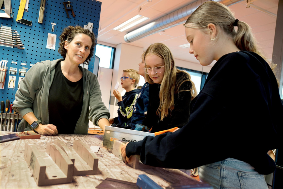 Vriendelijke dame met groenig overhemd over zwart t-shirt kijkt in de lens terwijl ze leerlingen begeleidt.