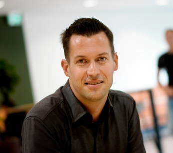 Ontwerp Man met kort donker haar en donker overhemd kijkt glimlachend in de camera in een binnenruimte, met twee personen wazig op de achtergrond.zonder titel (6)