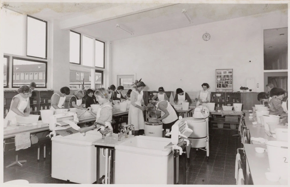 Een zwart-wit foto uit de jaren '60 waar een wasles is te zien in het waslokaal van de Eerste Openbare Huishoudschool. Op de foto staan maar twee wasmachines. Tijdens de lessen leerden de meeste leerlingen nog steeds hoe je met de hand wast - beeld bij verhaal over de huishoudschool op AOb.nl/Onderwijsblad