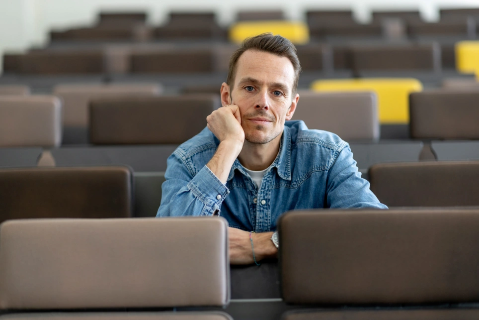 Foto van een jonge man in spijkerblouse in een lege collegezaal.
